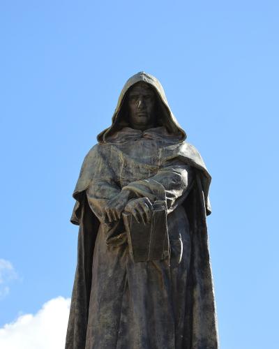 A bronze statue of a hooded figure holding a book, set against a blue sky with clouds, identified by the filename as Giordano Bruno in Campo de' Fiori.