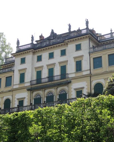 A large, multi-story neoclassical villa with pale yellow stucco walls, green shutters, and statues lining the roofline, surrounded by lush green hedges and trees under a bright sky.