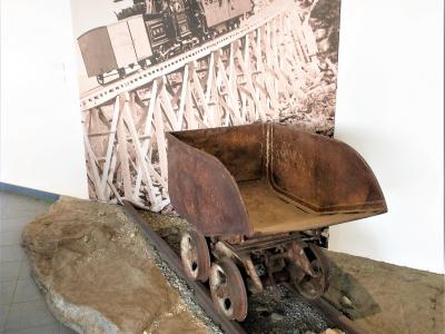 An indoor museum exhibit featuring a rusty, vintage mining or railway cart on a short section of track set into a simulated rocky base, with a large black-and-white historical photograph of a steam train on a trestle bridge mounted on the wall beh