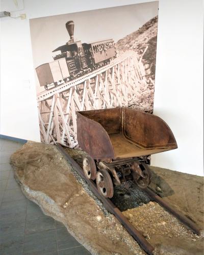 An indoor museum exhibit featuring a rusty, vintage mining or railway cart on a short section of track set into a simulated rocky base, with a large black-and-white historical photograph of a steam train on a trestle bridge mounted on the wall beh