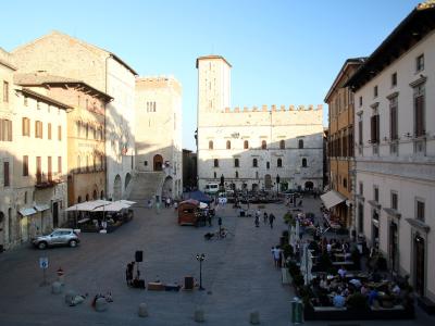 A low-angle photograph of a large, medieval stone building with crenellated battlements, arched windows, and an arched passageway on the left, situated in a paved public square under a blue sky.