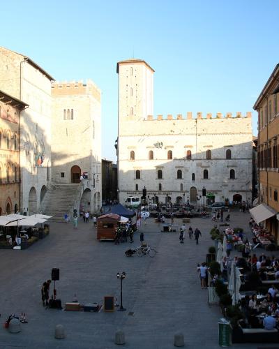 A low-angle photograph of a large, medieval stone building with crenellated battlements, arched windows, and an arched passageway on the left, situated in a paved public square under a blue sky.