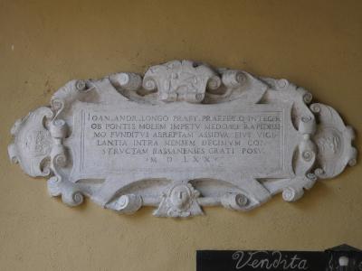 A close-up of an ornate stone plaque mounted on a yellow wall, featuring Latin text that includes "BASSANENSES" and the date "M D LXX" (1570), with a small black sign reading "Vendita" below it.