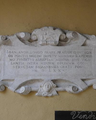 A close-up of an ornate stone plaque mounted on a yellow wall, featuring Latin text that includes "BASSANENSES" and the date "M D LXX" (1570), with a small black sign reading "Vendita" below it.