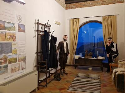 The image shows an indoor museum diorama featuring two life-sized mannequins in traditional attire, a coat rack, a window with a painted backdrop, and a wall display titled "Magna via Francigena" with informational panels and photos.