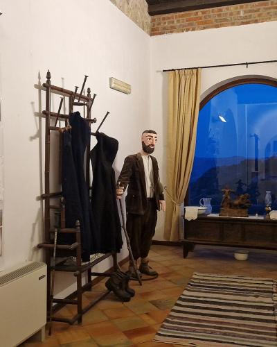The image shows an indoor museum diorama featuring two life-sized mannequins in traditional attire, a coat rack, a window with a painted backdrop, and a wall display titled "Magna via Francigena" with informational panels and photos.