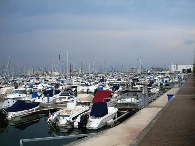 A crowded marina filled with numerous small motorboats and sailboats docked along a long concrete pier under an overcast sky, with industrial buildings and cranes visible in the background.