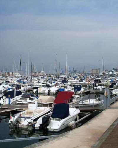 A crowded marina filled with numerous small motorboats and sailboats docked along a long concrete pier under an overcast sky, with industrial buildings and cranes visible in the background.