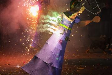 A person in a purple costume with fireworks attached dances at night during a festival, with sparks flying and another figure visible in the background.