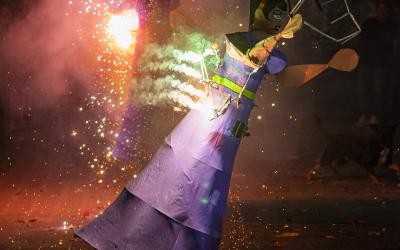 A person in a purple costume with fireworks attached dances at night during a festival, with sparks flying and another figure visible in the background.
