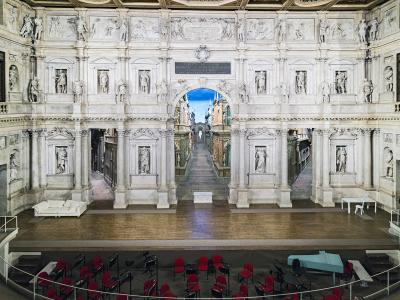 The image shows the interior of a Renaissance theater with an ornate, multi-tiered white stone facade featuring numerous statues, a central archway revealing a painted perspective scene of a street, and a wooden stage with red audience seating bel