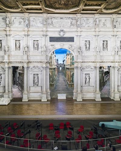 The image shows the interior of a Renaissance theater with an ornate, multi-tiered white stone facade featuring numerous statues, a central archway revealing a painted perspective scene of a street, and a wooden stage with red audience seating bel