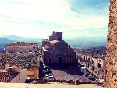 A high-angle view of a large stone church with a central dome and bell tower situated in a town square, surrounded by terracotta-roofed buildings and hills in the background.