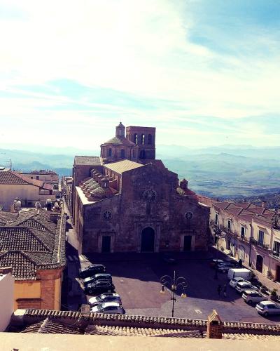 A high-angle view of a large stone church with a central dome and bell tower situated in a town square, surrounded by terracotta-roofed buildings and hills in the background.
