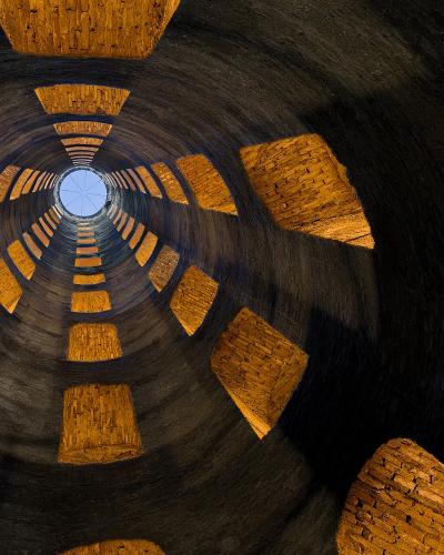 A view looking straight up the interior of a deep, cylindrical stone well with a double-helix ramp structure and a circular opening to the sky at the top.