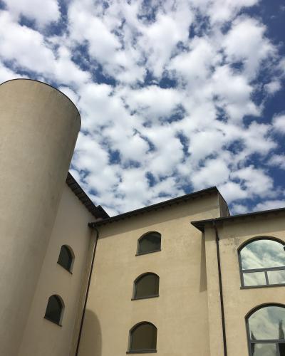 A low-angle view of a beige building with arched windows and a cylindrical tower against a cloudy blue sky.