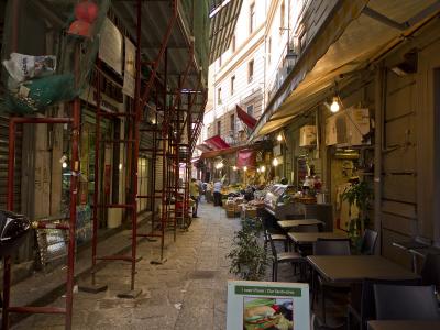 A narrow, paved alleyway in an urban setting, featuring red scaffolding on the left, outdoor dining tables on the right, and a sandwich shop sign reading "I nostri Panini / Our Sandwiches".