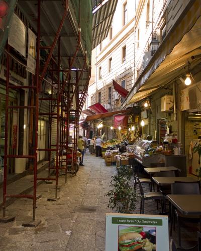 A narrow, paved alleyway in an urban setting, featuring red scaffolding on the left, outdoor dining tables on the right, and a sandwich shop sign reading "I nostri Panini / Our Sandwiches".