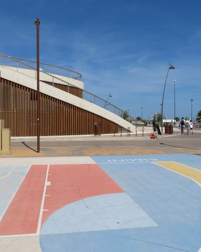 The image shows an outdoor basketball court with colorful painted surfaces, a Red Bull-branded hoop, and a modern wooden-and-concrete structure in the background; the word “RIMINI” is painted on the court surface.