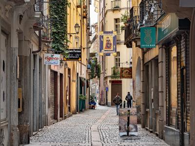 A narrow, cobblestone pedestrian street lined with historic buildings, featuring hanging signs for businesses like "Juice Bar" and "Cigolò," with two people walking in the distance.