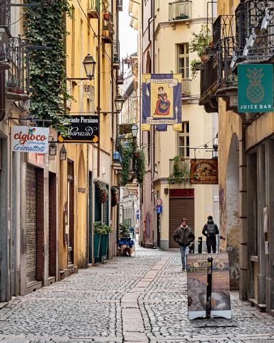 A narrow, cobblestone pedestrian street lined with historic buildings, featuring hanging signs for businesses like "Juice Bar" and "Cigolò," with two people walking in the distance.