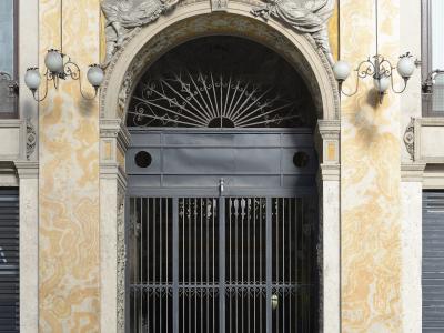 A close-up of an ornate arched entrance with a black metal gate, flanked by yellow marble-patterned walls and topped with sculpted winged figures and the number "27" on a plaque.