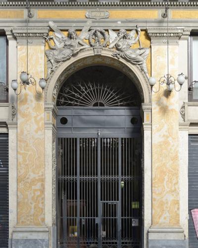 A close-up of an ornate arched entrance with a black metal gate, flanked by yellow marble-patterned walls and topped with sculpted winged figures and the number "27" on a plaque.