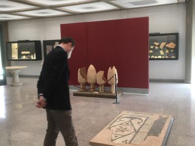 A man in a dark jacket and face mask walks through a modern museum gallery featuring ancient artifacts, including a large floor mosaic on a platform in the foreground and several standing stone fragments against a red partition wall.