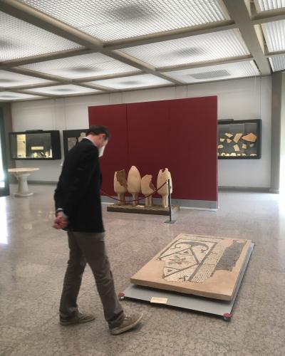 A man in a dark jacket and face mask walks through a modern museum gallery featuring ancient artifacts, including a large floor mosaic on a platform in the foreground and several standing stone fragments against a red partition wall.