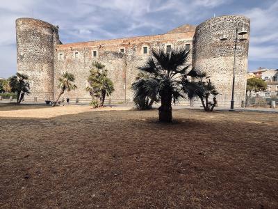 A low-angle exterior shot of a large medieval stone fortress with two prominent cylindrical corner towers, situated behind a dry grassy lawn with several palm trees under a partly cloudy sky.