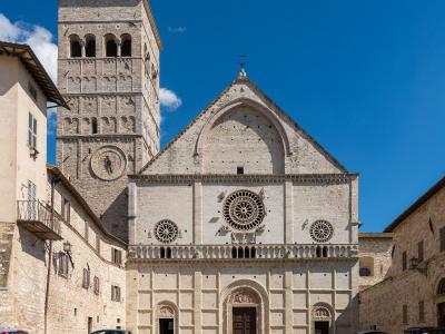 The image shows the facade and bell tower of a large Romanesque stone cathedral with a prominent rose window, three arched doorways, and a square bell tower featuring a sundial, set against a clear blue sky in a paved plaza with parked cars.