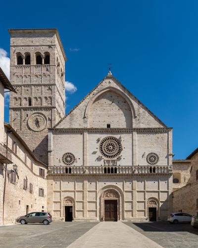 The image shows the facade and bell tower of a large Romanesque stone cathedral with a prominent rose window, three arched doorways, and a square bell tower featuring a sundial, set against a clear blue sky in a paved plaza with parked cars.