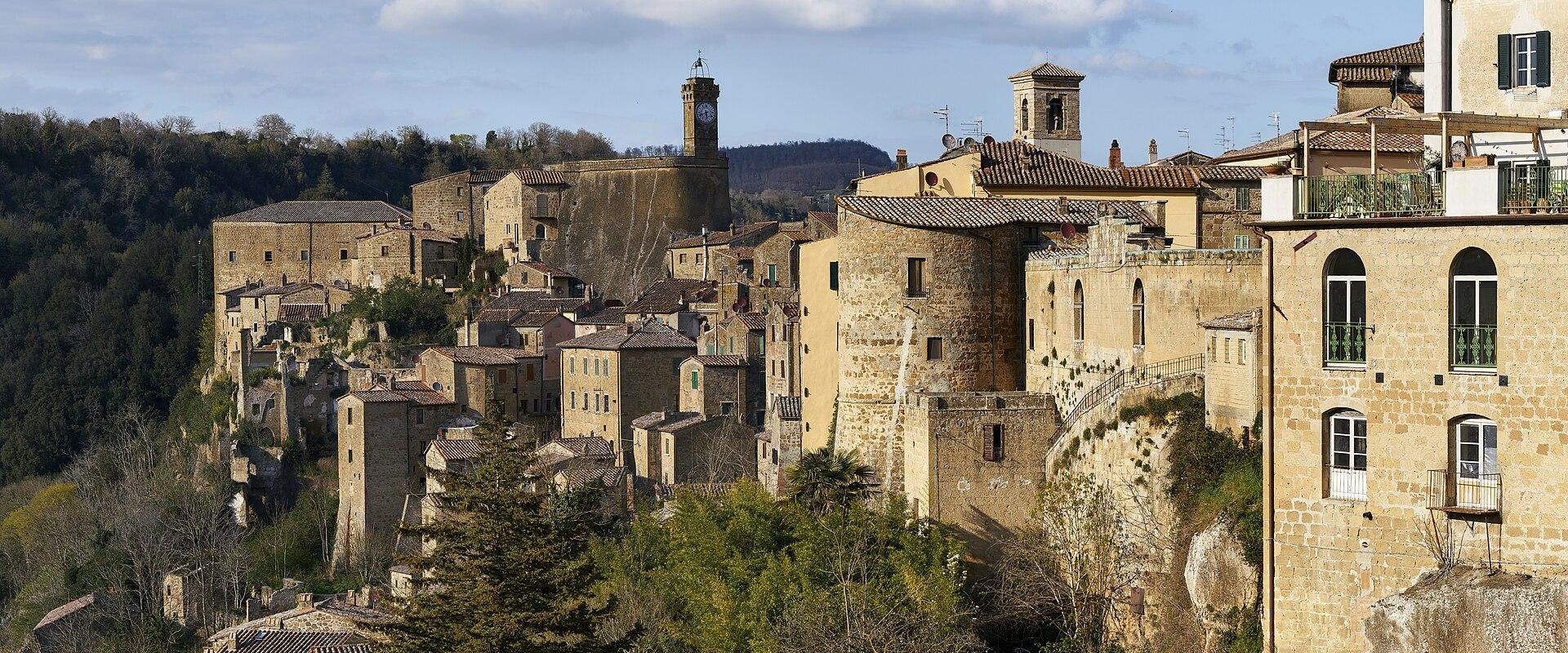A panoramic view of the historic hilltop village of Sorano, featuring stone buildings, a prominent clock tower, and terracotta roofs set against a backdrop of rolling hills and a partly cloudy sky.