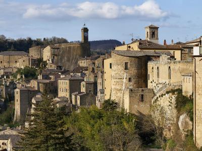 A panoramic view of the historic hilltop village of Sorano, featuring stone buildings, a prominent clock tower, and terracotta roofs set against a backdrop of rolling hills and a partly cloudy sky.
