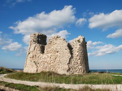 A stone ruin of a coastal tower with three distinct sections, set on a grassy dune with the sea in the background under a blue sky.