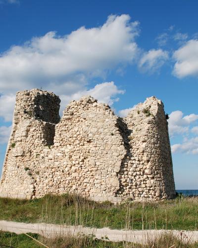 A stone ruin of a coastal tower with three distinct sections, set on a grassy dune with the sea in the background under a blue sky.