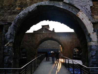 The image shows a large, ancient stone archway with visible weathering and layered stonework, framing a pedestrian walkway with modern metal railings; people are walking through the arch under bright sunlight, with buildings visible in the backgro