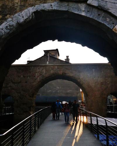 The image shows a large, ancient stone archway with visible weathering and layered stonework, framing a pedestrian walkway with modern metal railings; people are walking through the arch under bright sunlight, with buildings visible in the backgro