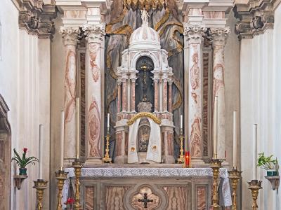 The image shows an ornate interior chapel altar with marble columns, a gilded canopy, religious statuary, candles, and potted plants, featuring a checkered floor and rope barriers.