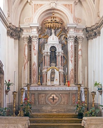The image shows an ornate interior chapel altar with marble columns, a gilded canopy, religious statuary, candles, and potted plants, featuring a checkered floor and rope barriers.