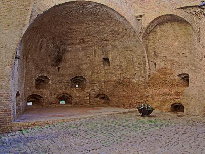 The image shows the interior of a large, vaulted brick structure with arched openings and a cobblestone floor, featuring a central planter and a wall-mounted lamp; the architecture suggests a historic defensive or utilitarian space like a gatehous