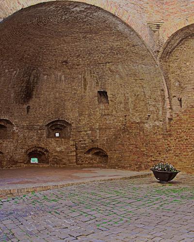The image shows the interior of a large, vaulted brick structure with arched openings and a cobblestone floor, featuring a central planter and a wall-mounted lamp; the architecture suggests a historic defensive or utilitarian space like a gatehous