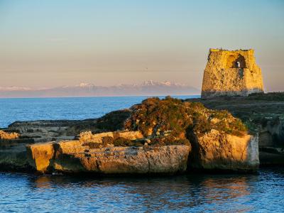A stone coastal tower ruin on a rocky promontory overlooking the sea, with snow-capped mountains visible in the distant background under a clear sky.