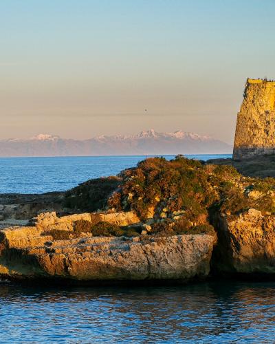 A stone coastal tower ruin on a rocky promontory overlooking the sea, with snow-capped mountains visible in the distant background under a clear sky.