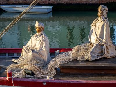 Two bronze statues of fishermen sitting on the deck of a wooden boat with fishing nets, located in a canal or harbor setting.