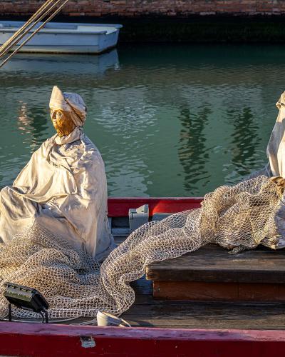 Two bronze statues of fishermen sitting on the deck of a wooden boat with fishing nets, located in a canal or harbor setting.