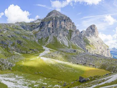 A panoramic landscape photograph showing a prominent, rugged mountain peak rising above green alpine meadows and scree slopes under a blue sky with scattered clouds; no text, signage, or map elements are visible in the image itself.