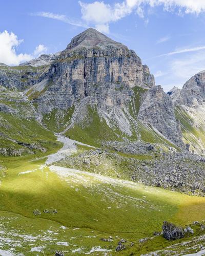 A panoramic landscape photograph showing a prominent, rugged mountain peak rising above green alpine meadows and scree slopes under a blue sky with scattered clouds; no text, signage, or map elements are visible in the image itself.