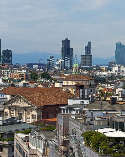 A panoramic cityscape showing a dense urban foreground of terracotta-roofed buildings and historic architecture, transitioning to a modern skyline of glass skyscrapers in the distance, with mountains visible on the horizon under a partly cloudy sk