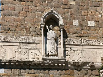 A close-up of a stone wall featuring a Gothic-style niche with a statue of a robed figure holding a palm frond, flanked by carved reliefs and set against a backdrop of aged masonry under a clear blue sky.